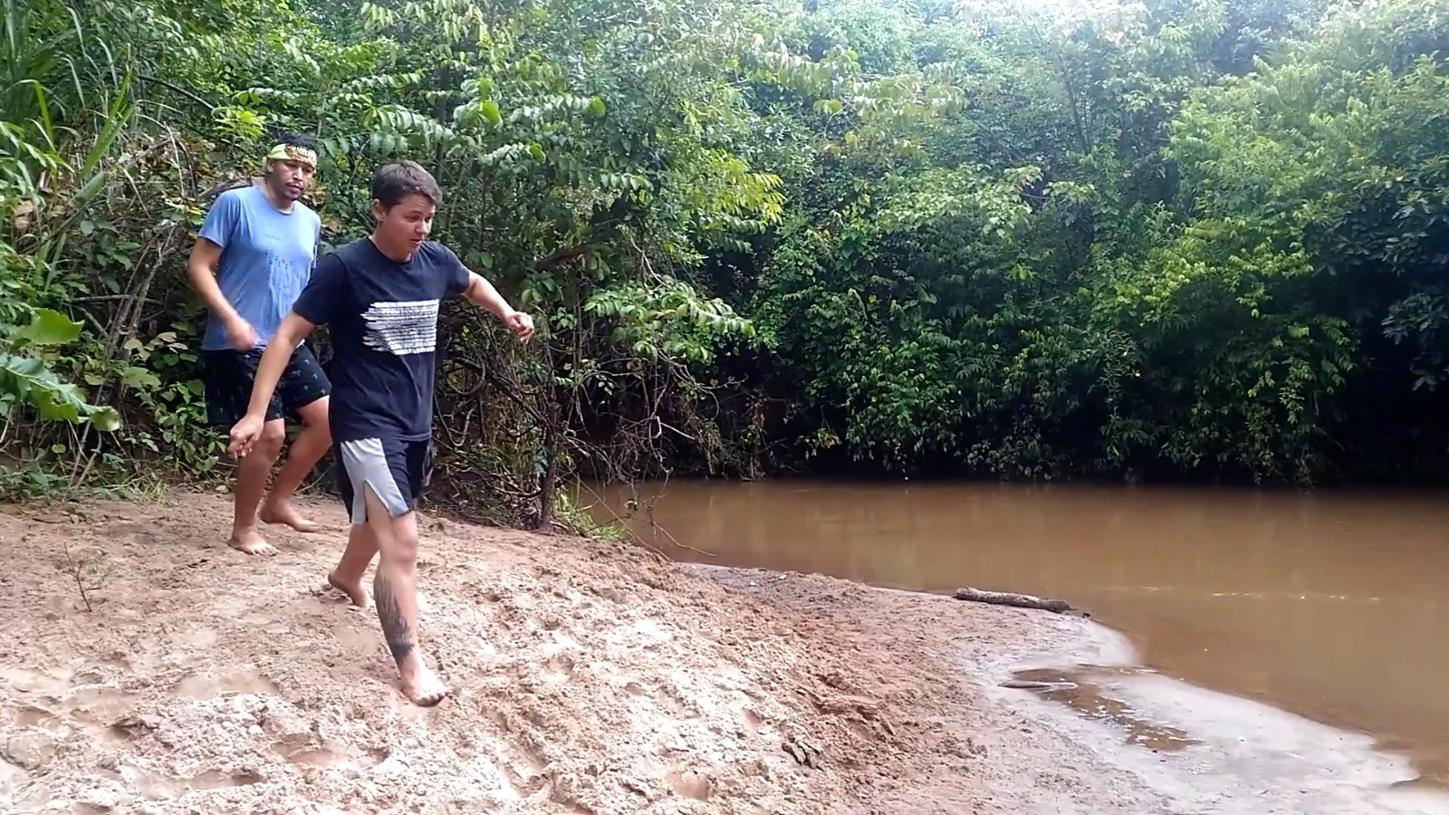 Duas pessoas caminhando descalças em uma margem de rio com areia, cercadas por vegetação densa e árvores, em um ambiente natural.