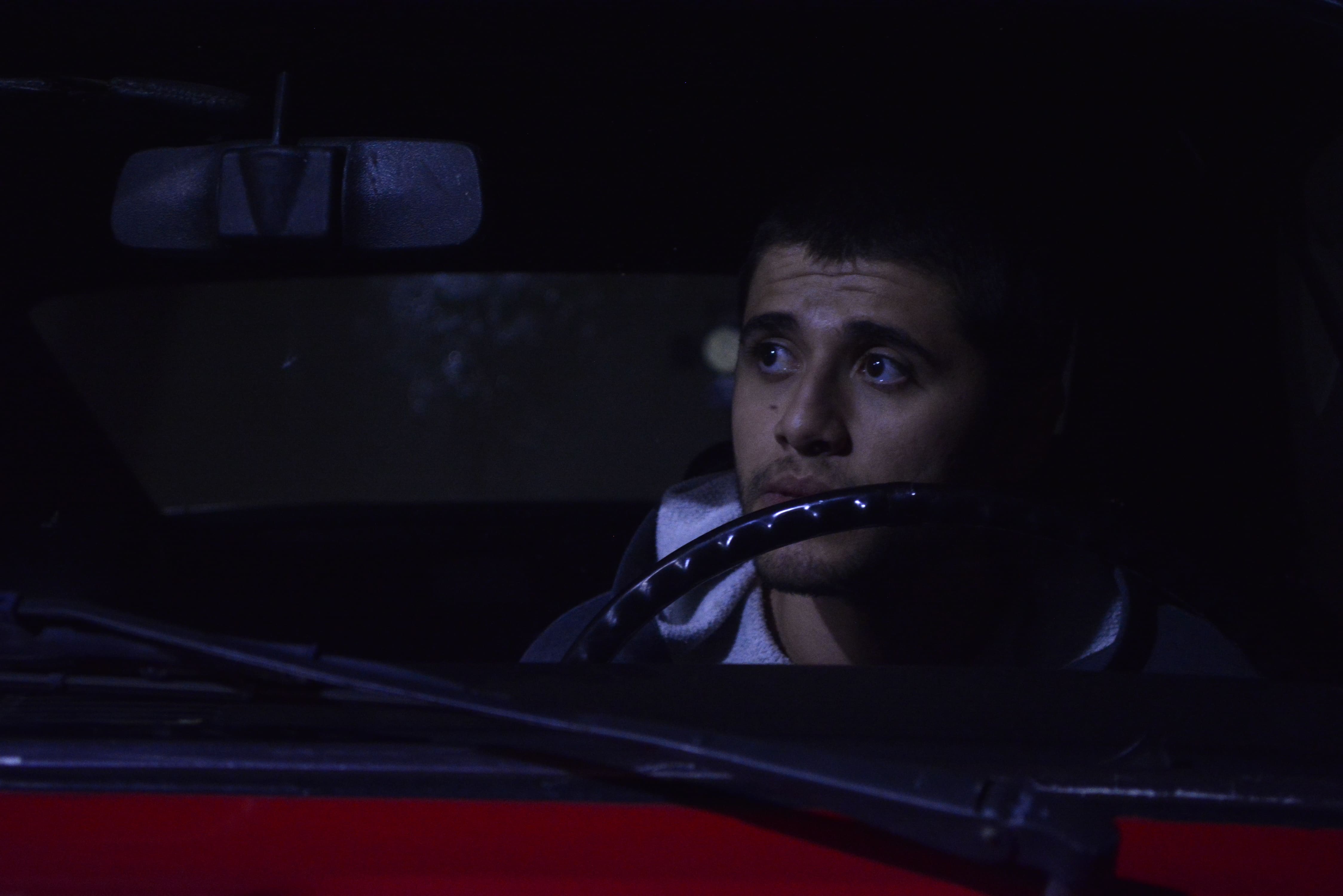 Homem jovem dentro de um carro à noite, olhando pensativo para fora com expressão séria, iluminado por uma luz suave. A imagem é escura, com destaque para o rosto do rapaz e o volante à sua frente.