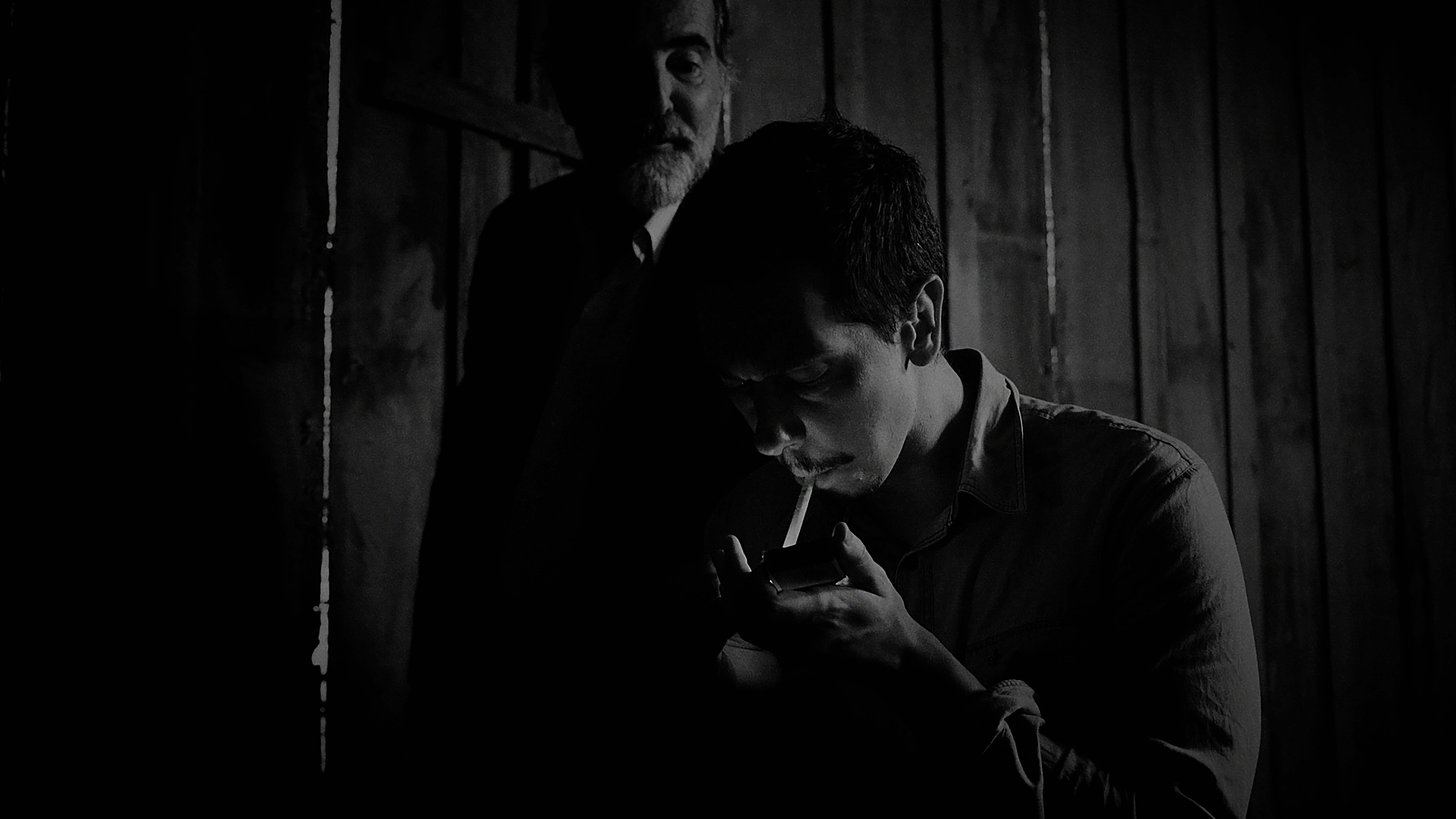 Imagem em preto e branco de dois homens em uma sala escura. Um homem ao fundo, com barba e expressão séria, observa enquanto o homem em primeiro plano está concentrado em acender um cigarro com um isqueiro. A iluminação fraca cria um efeito dramático com sombras intensas.