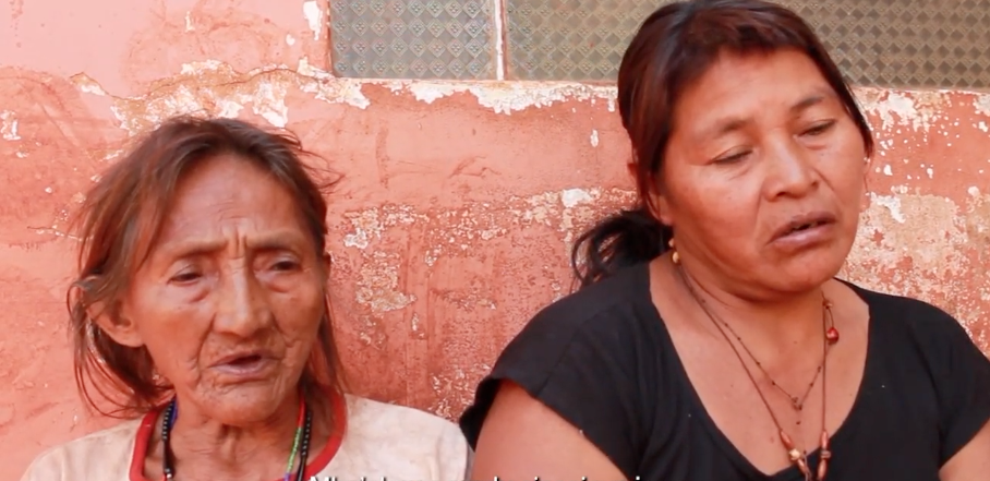 Duas mulheres sentadas contra uma parede avermelhada e desgastada, uma idosa com pele enrugada e usando um colar tradicional, e a outra mais jovem de olhos fechados, vestindo uma camiseta escura e um colar com pingente, ambas aparentando estar em uma conversa ou reflexão profunda.