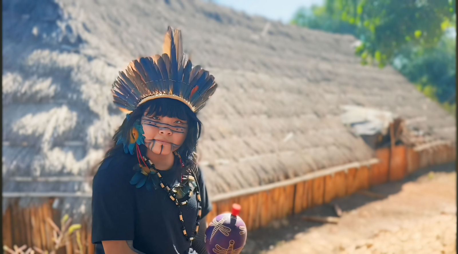Criança indígena com cocar de penas, pintura facial tradicional e segurando um instrumento pintado, em frente a uma construção com telhado de palha em ambiente rural.